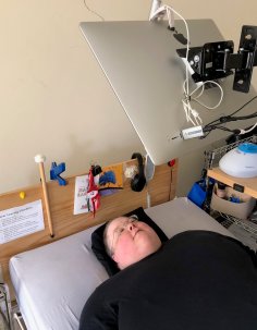 Woman is laying on her bed looking up.  There is a computer screen mounted above her head that she is viewing.