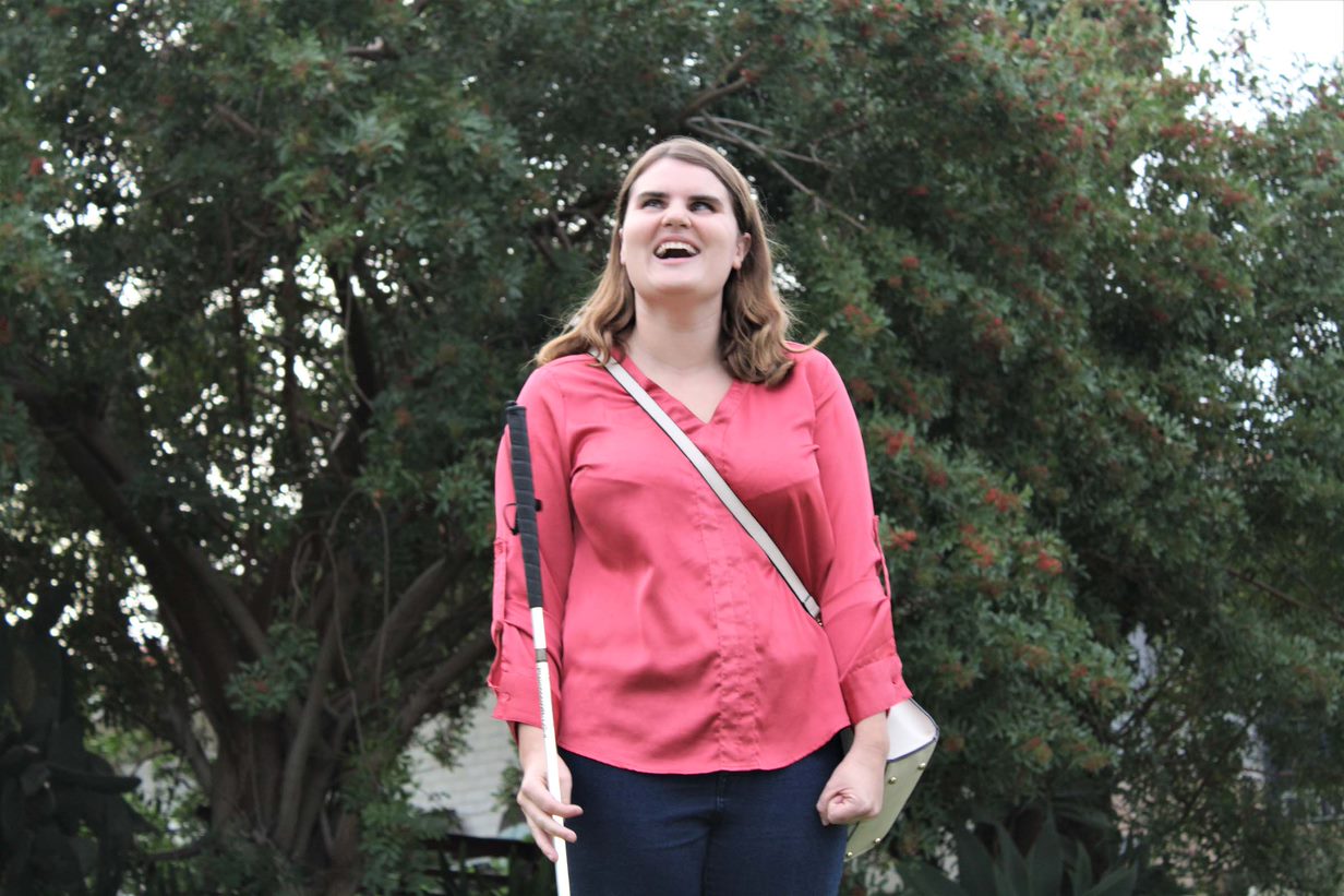 A woman stands in front of her house and holds her white cane, smiling. She stands in front of a garden..