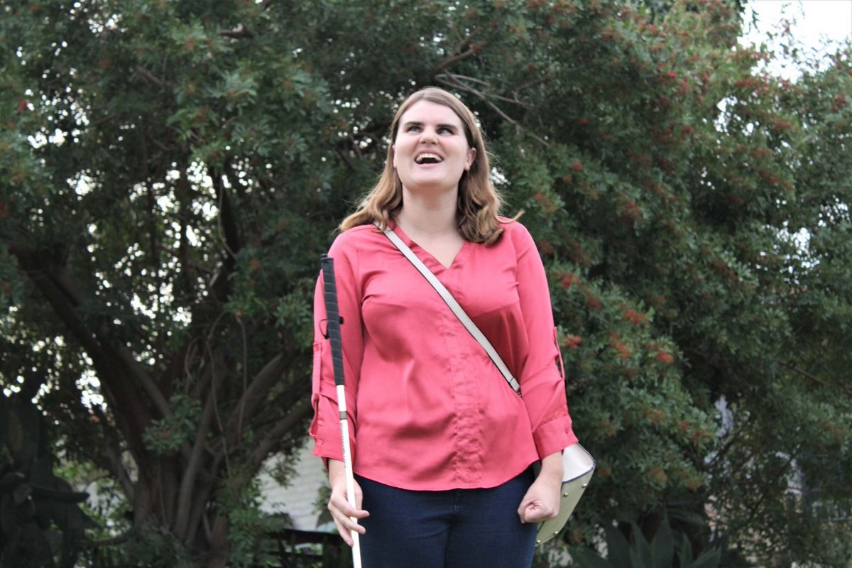 A lady stand on the lawn smiling as she holds her white cane