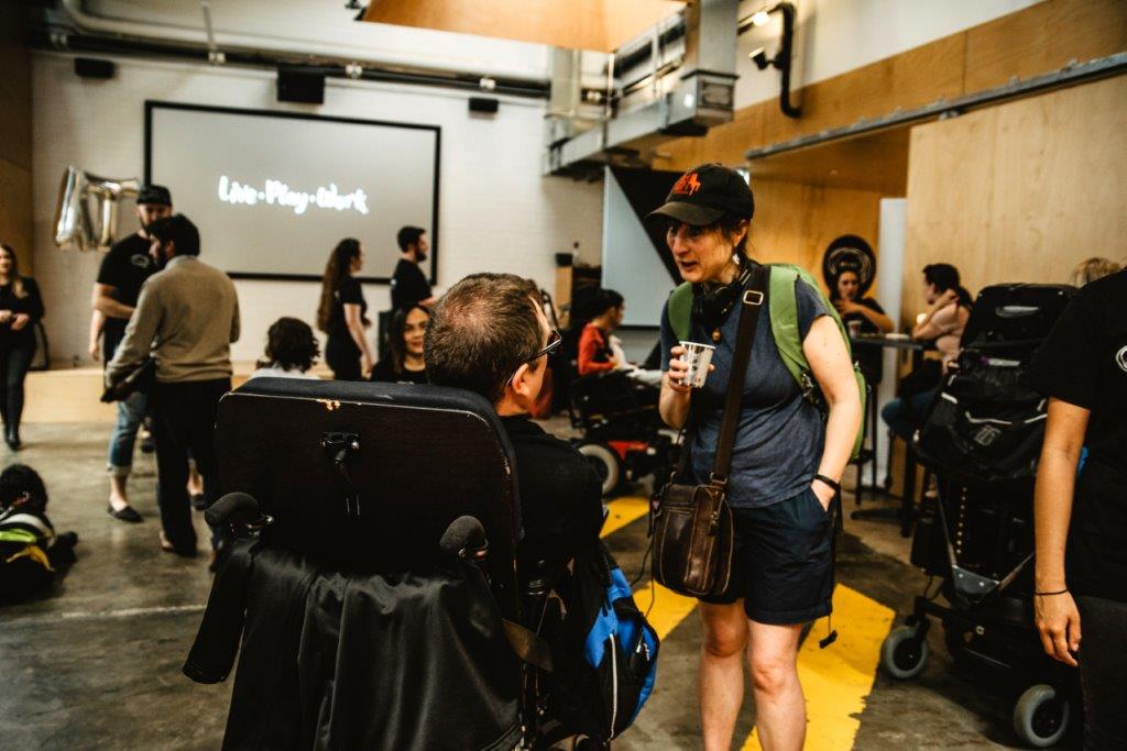 Two people are chatting at the AT Chat launch.  One is seated in a power wheelchair