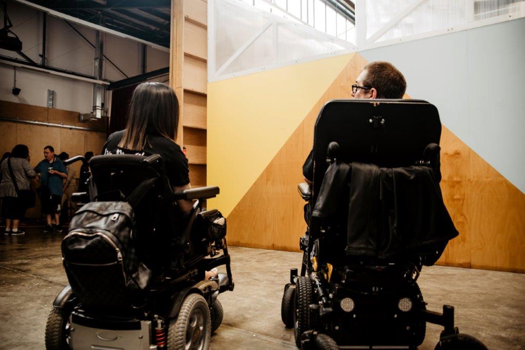Back of two people using power wheelchairs.  They are seated in front of a brown wall