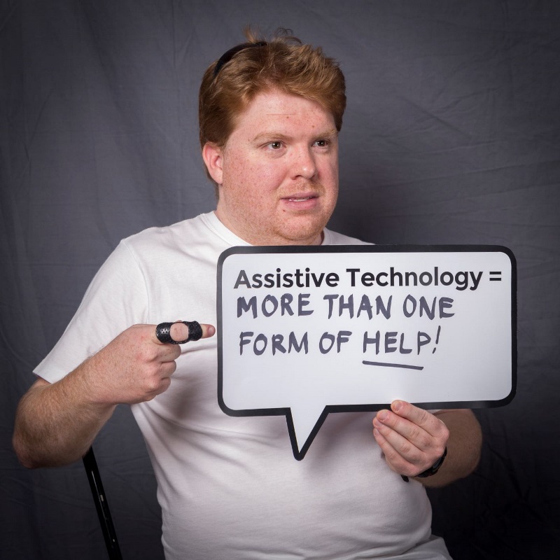 chris_speechbubble young man holds up speech bubble sign that reads assistitve technology = more than one form of help!