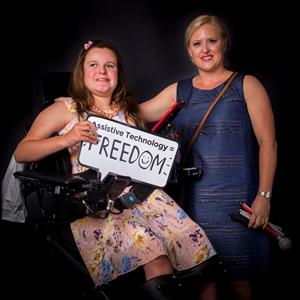 a woman wearing a blue dress stands next to her daughter who is sitting in a elevated wheelchair holding a sign that reads assistive technology = freedom
