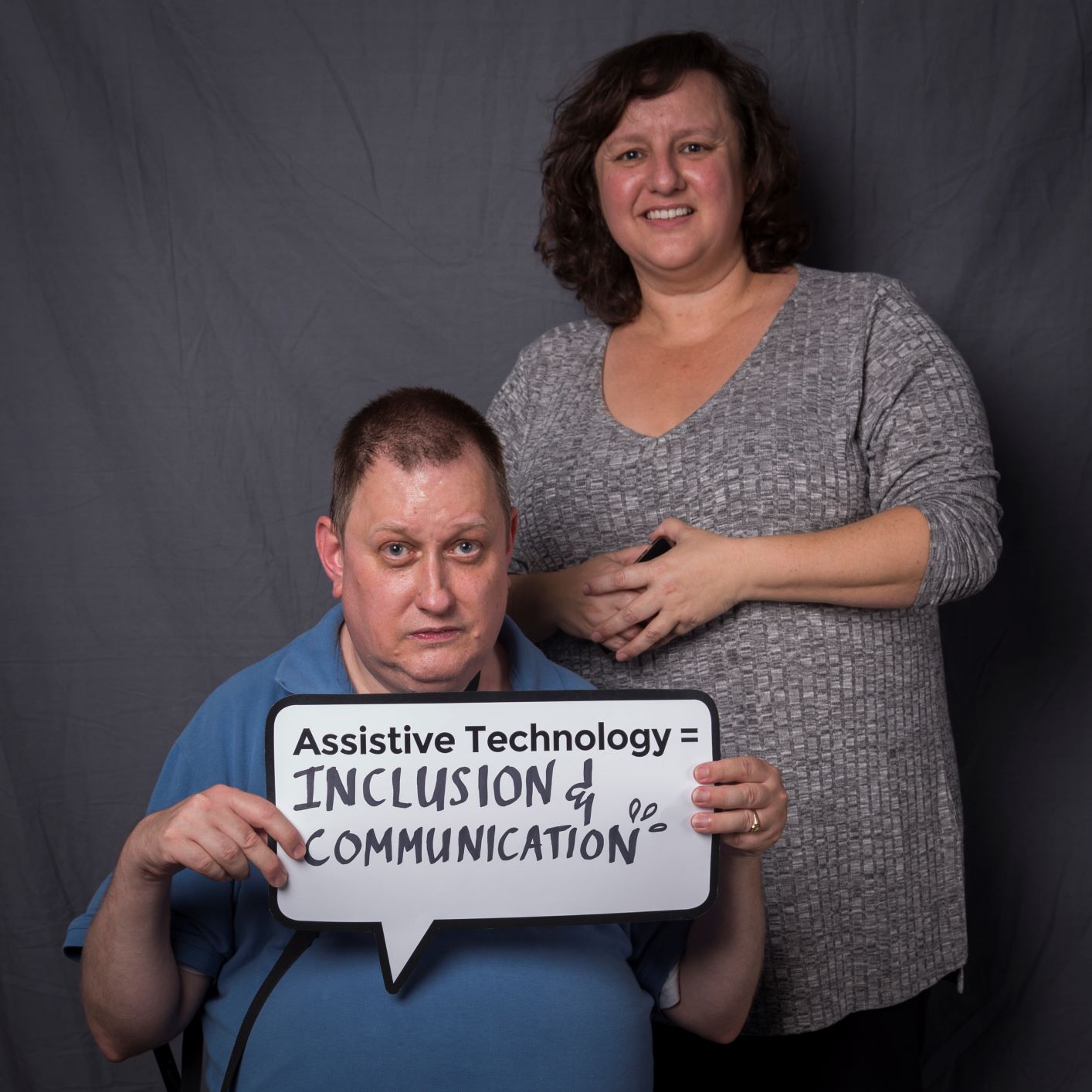 a woman stands behind a seated man holding a speech bubble sign that reads assistive technology = inclusion and communication
