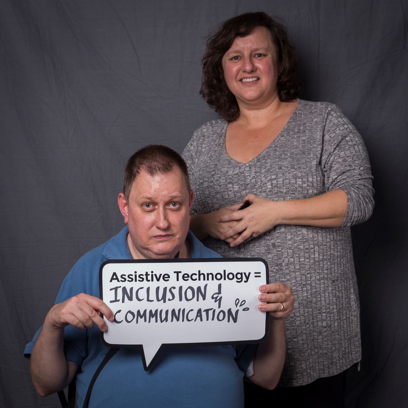 inclusion and communication a woman stands behind a seated man holding a speech bubble sign that reads assistive technology = inclusion and communication