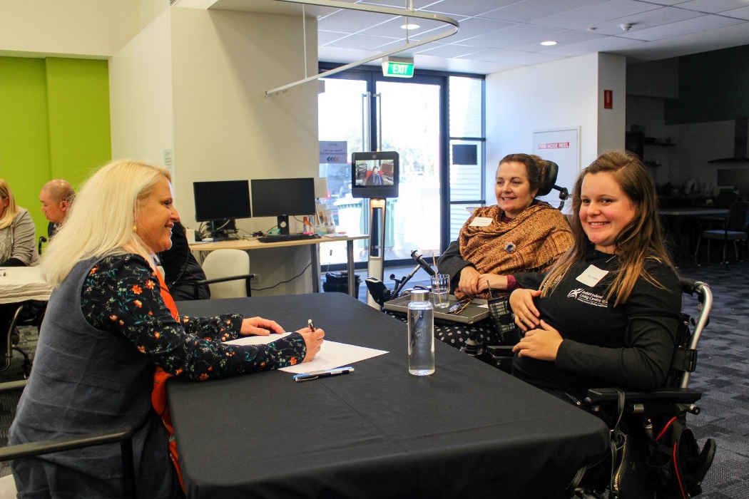 three woman sit around a table with another woman shown on a video call on a screen