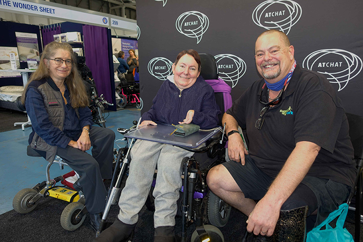 Two women and a man are seated beside each other in front of the AT Chat banner smiling at the camera. One is seated on her travel scooter, one in her powered wheelchair and the man on a seat with his prosthetic leg stretched out in front of him.