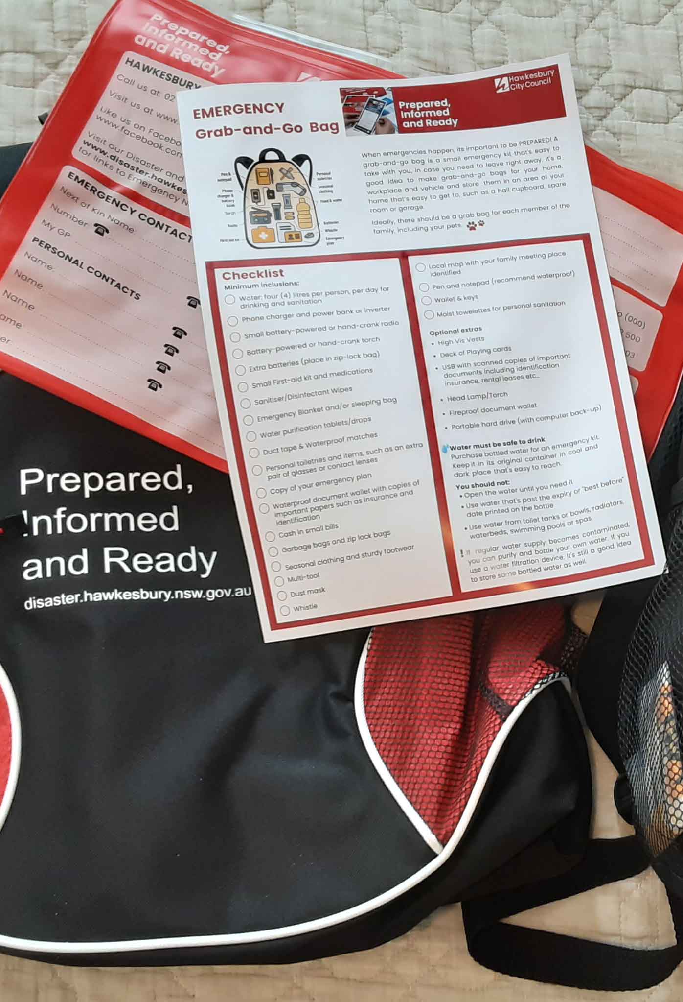 Grab-and-Go-Bag A red and black bag with the words, prepared, informed and ready stiched to the front along with the webaddress, disaster.hawksbury.nsw.gov.au. A emergency grab and go bag checklist sits on top of it.