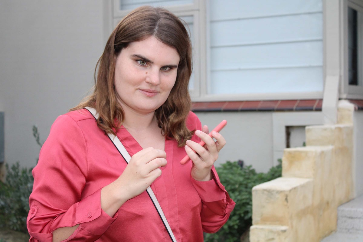 A woman stands in front of her house and holds her phone close to her face.