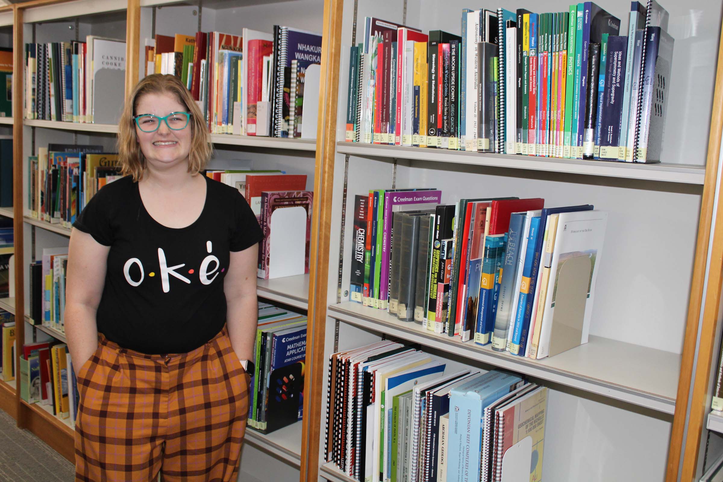 A lady smiles brightly and stands in front of bookshelves with her hands in her pockets.