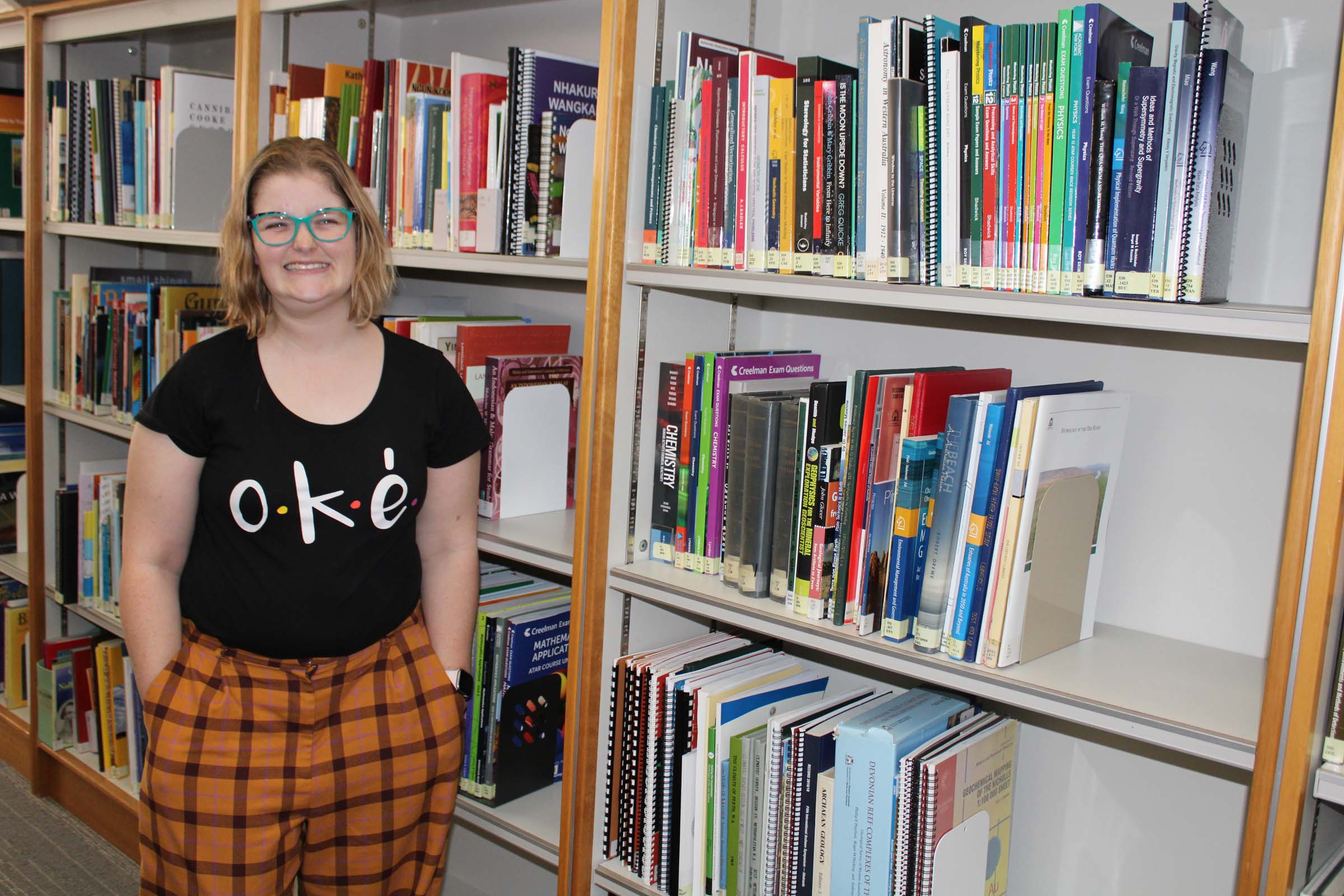 A lady smiles brightly and stands in front of bookshelves with her hands in her pockets. A lady smiles brightly and stands in front of bookshelves with her hands in her pockets.