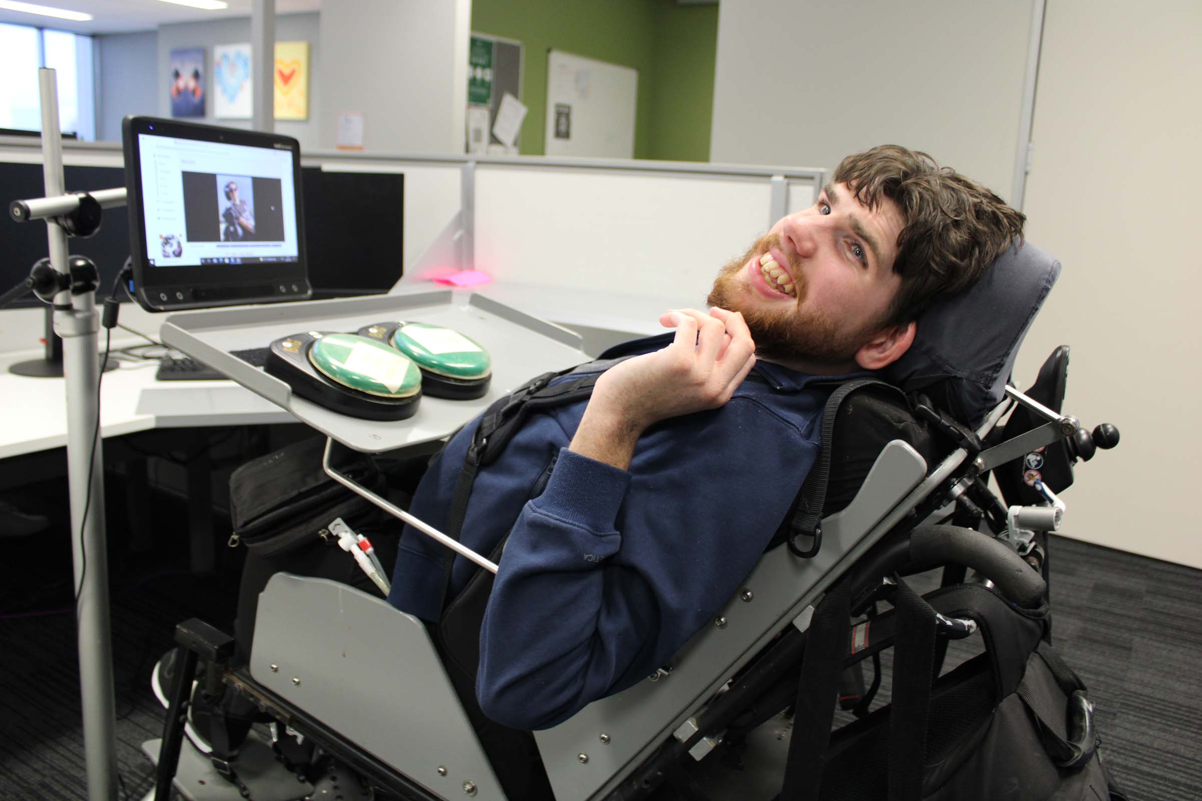 A man looks up from his powered wheelchair and smiles as he works at a computer screen. A man looks up from his powered wheelchair and smiles as he works at a computer screen.