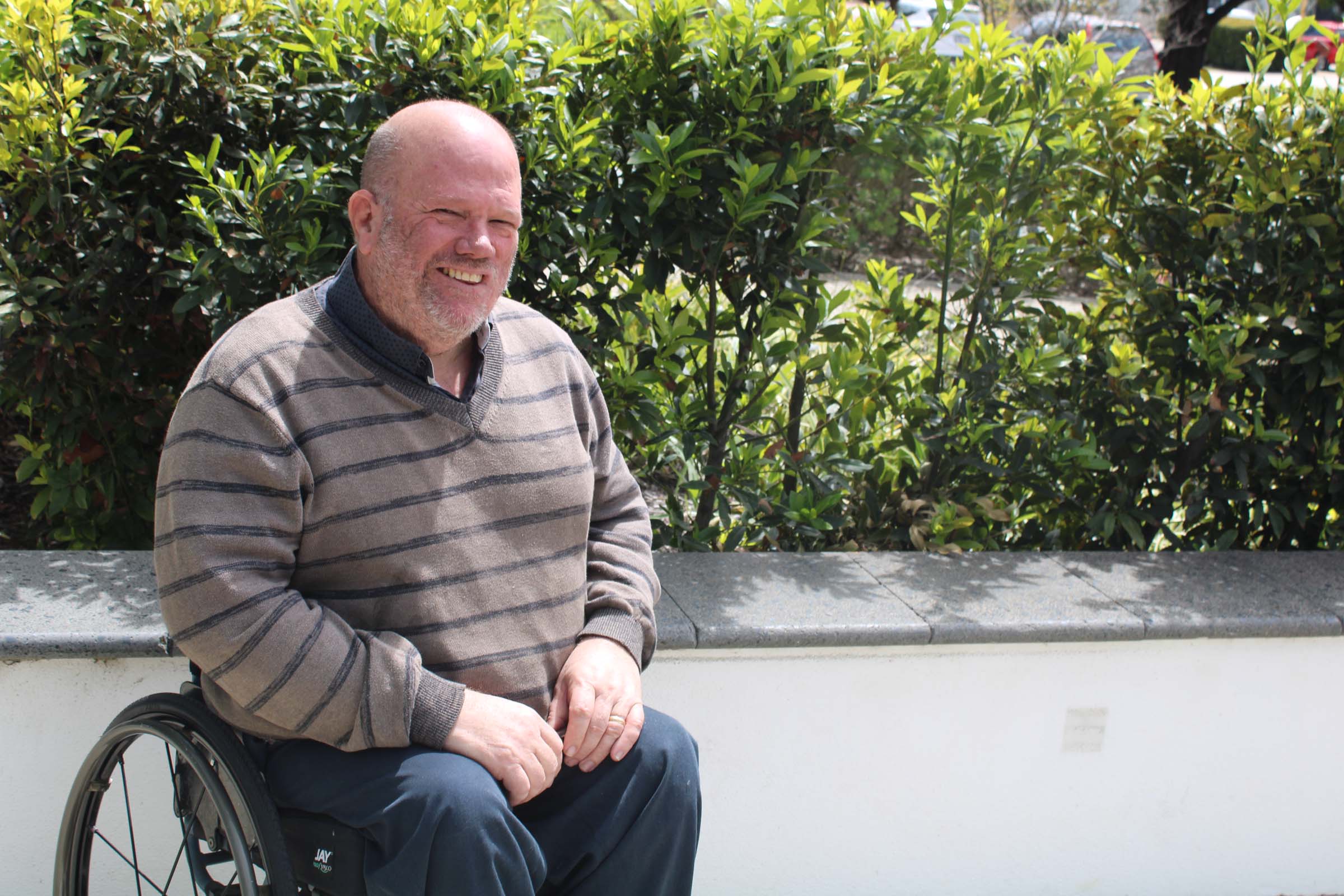 A man sits in a wheelchair next to a garden bed and smiles at the camera.