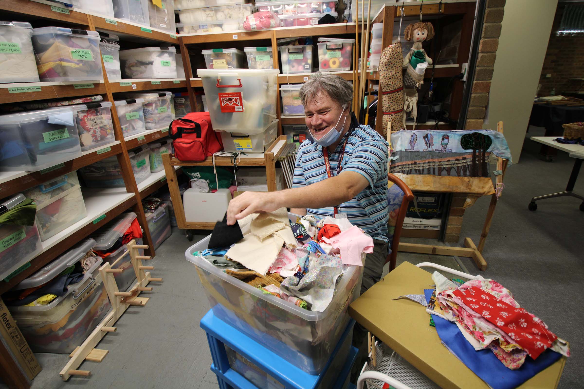 A man sits in a craft room, smiles and sorts through linen.