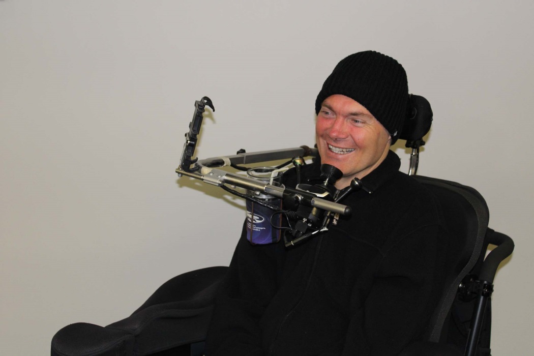 A man sits in his powered wheelchair and smiles. He wears a beanie on his head and a dark jumper.