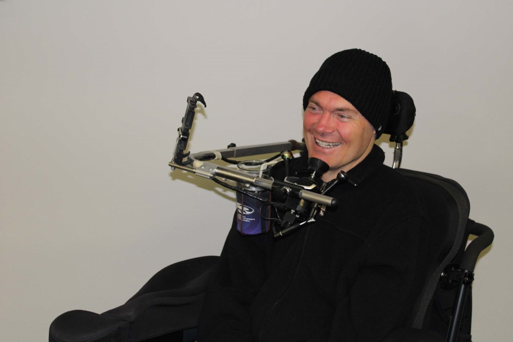 A man sits in his powered wheelchair and smiles. He wears a beanie on his head and a dark jumper. A man sits in his powered wheelchair and smiles. He wears a beanie on his head and a dark jumper.