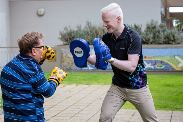 A man runs a training session with another man. They are wearing boxing gloves and smiling.