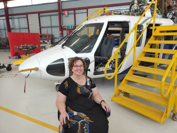 A lady sits in her wheelchair and smiles brightly. She is in an airplane hangar and a plane is behind her.