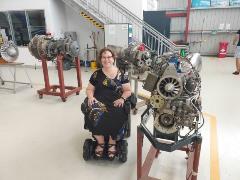 Nerine smiles and sits in her wheelchair next to a jet engine