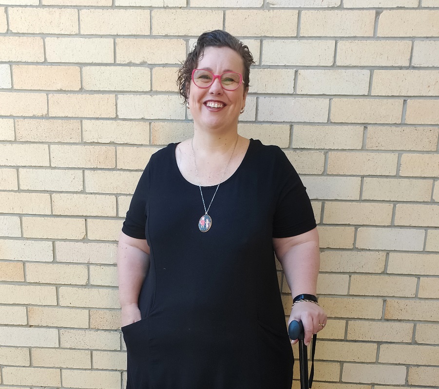 A lady stands in front of a pale brick wall holding her walking stick