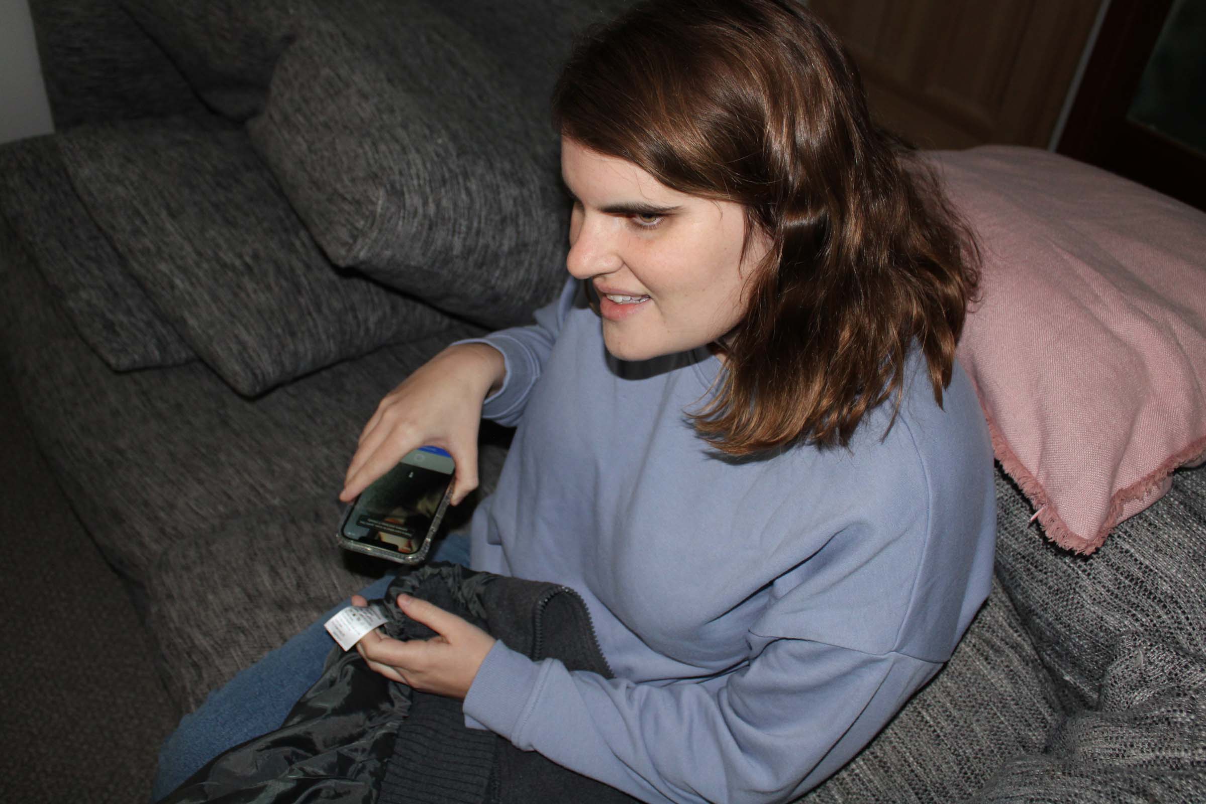 A woman sits on the couch and smiles, holding a smart phone.