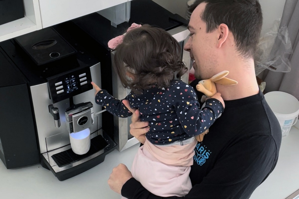 A man is standing in his kitchen making coffee using a commercial coffee machine, he holds a young girl in his arms.