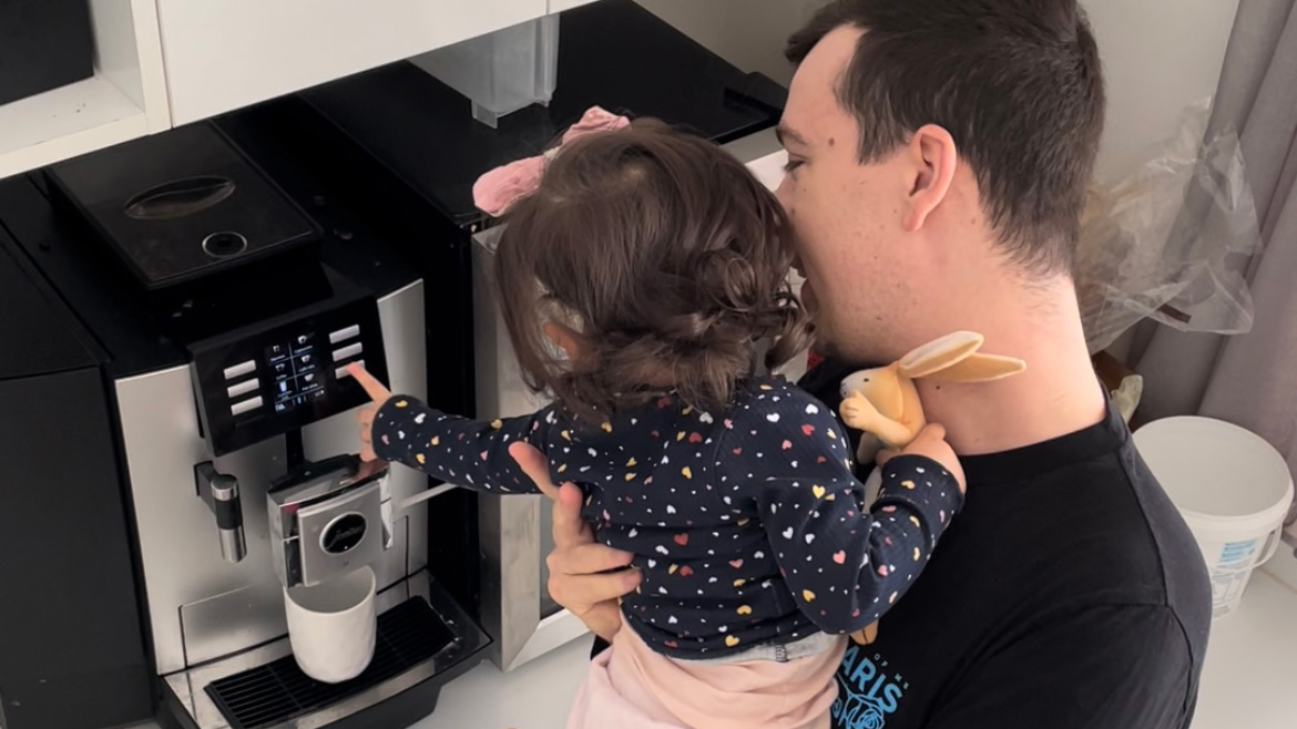 A man holds a young girl on his hip as she leans forward to push a button on a coffee machine