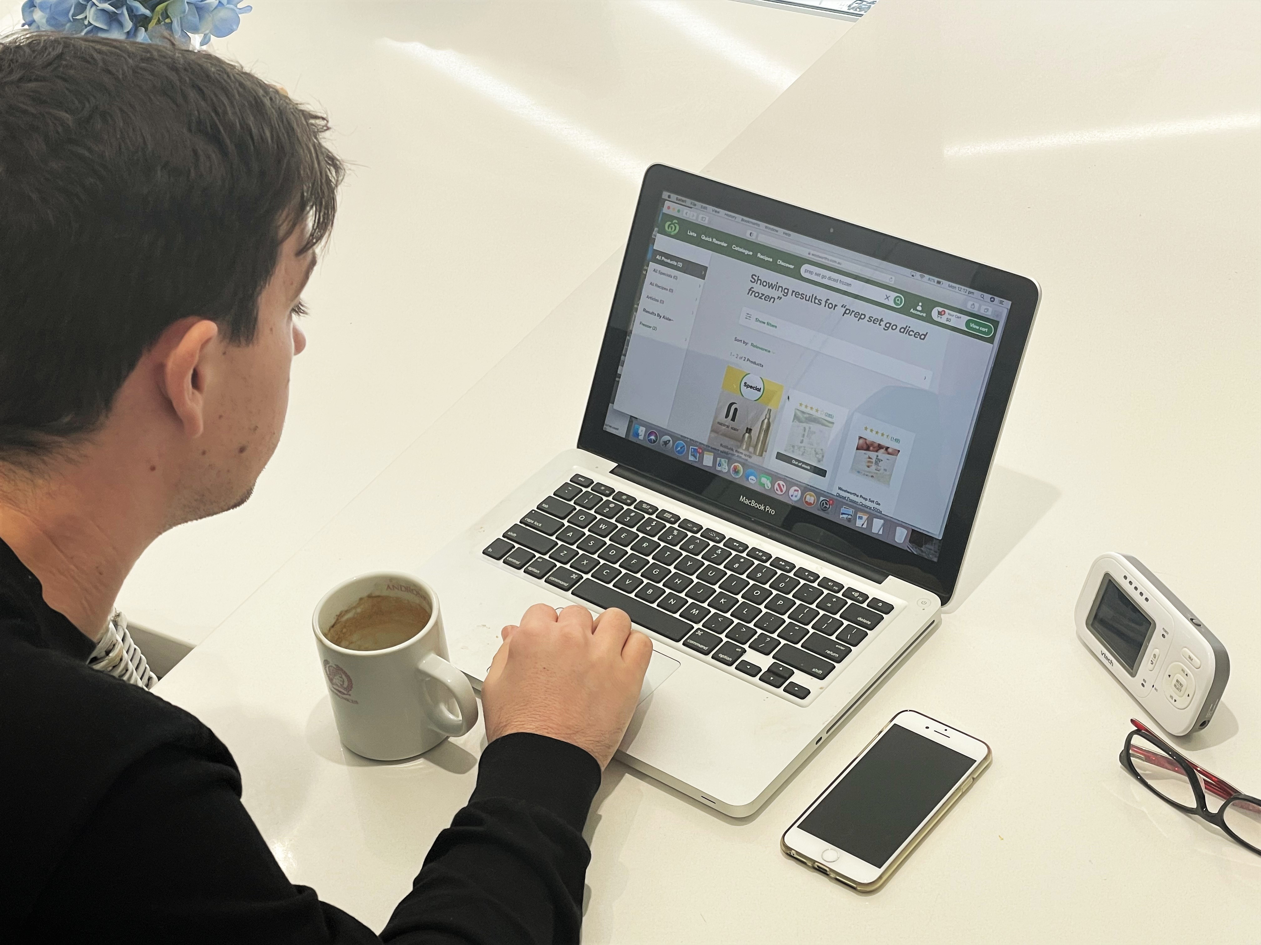 A man is using his laptop to make a purchase from Woolworths. He has his mobile phone, a timer, glasses and cup of coffee beside him.