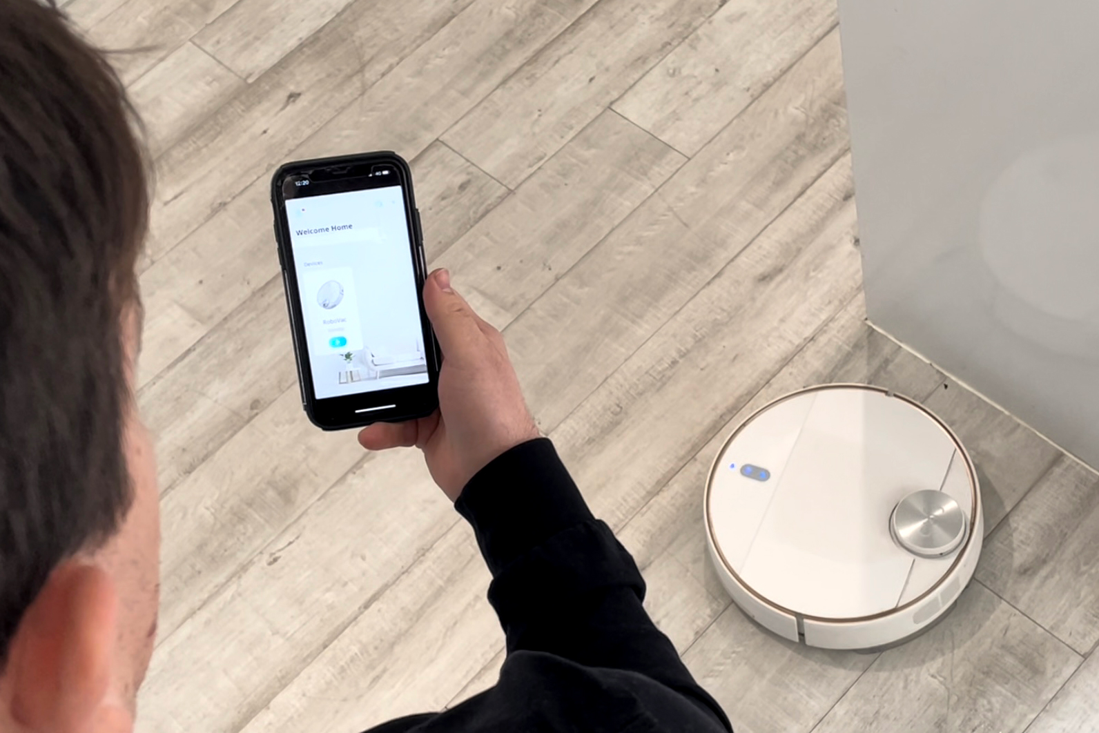 A man uses the Robovax app on his mobile phone. The Robovax vacuum is on the floor in front of him.