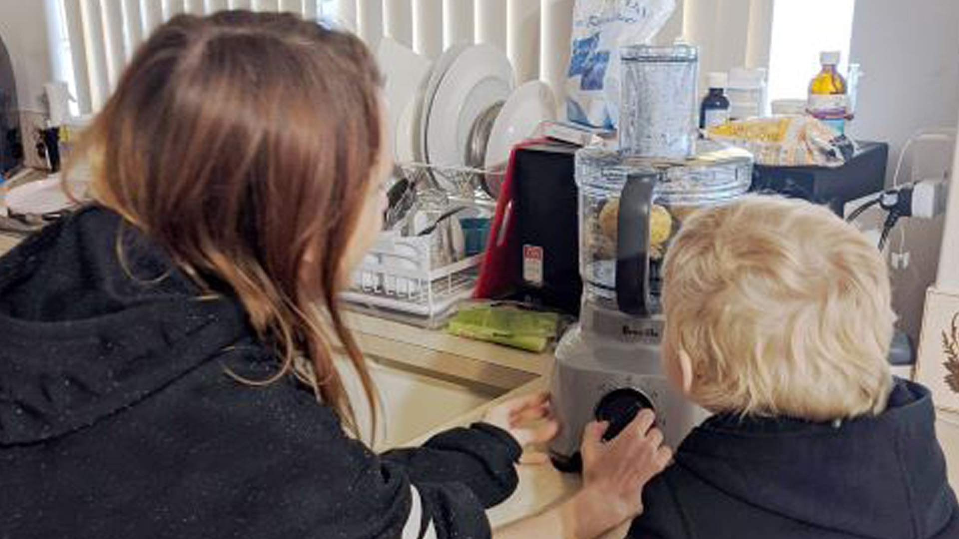 A woman and a young boy are leaning over a kitchen counter setting the dial on a kitchen wiz.