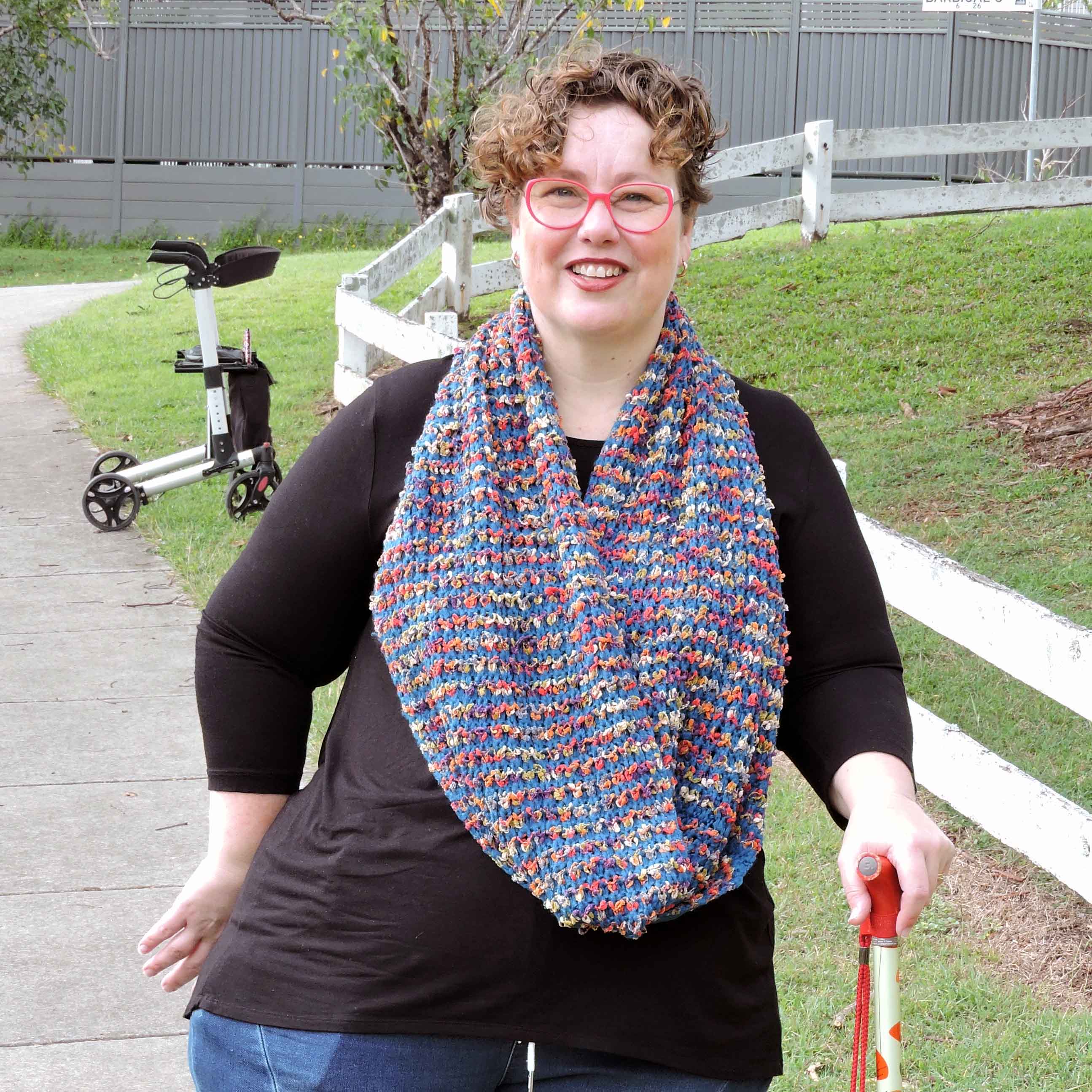 A woman is smiling as she walks along a footpath using her colourful walking stick. Her rollator is on the footpath behind her.