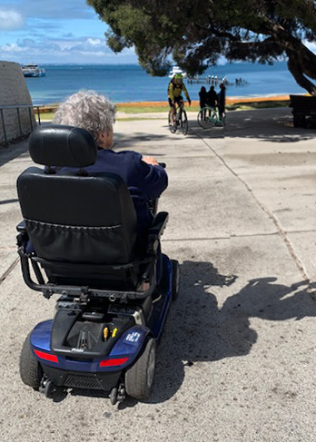 woman is seated in her mobility scooter with her back to the camera. She is looking out to sea watching people on bikes come towards her.
