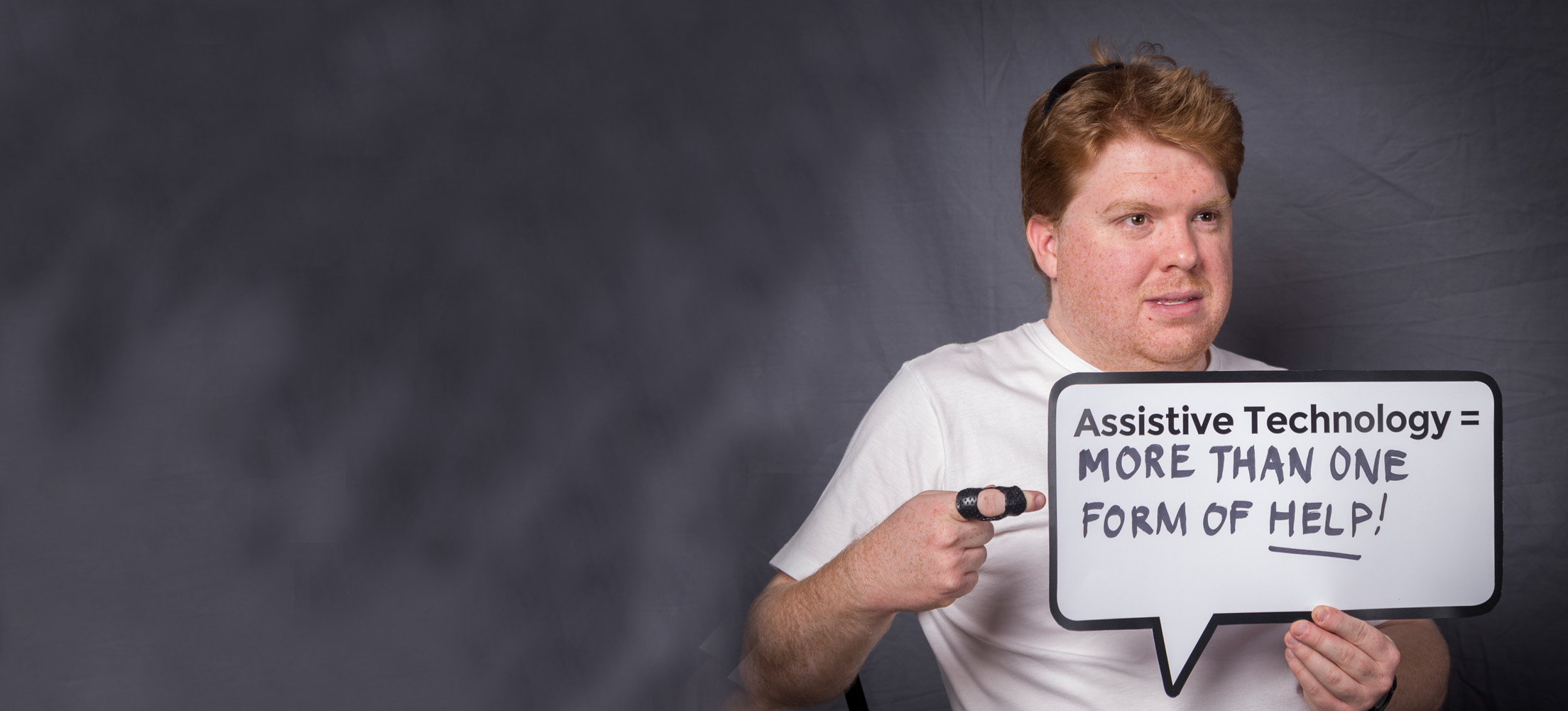A young man is pointing at a cardboard speech bubble that says, assistive technology equals more than one form of help.