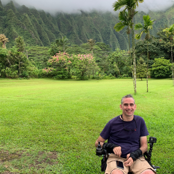 Photo of a man in a wheelchair in front of a scene of rainforest creeping up mountains with mist hanging over them