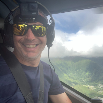 A man with a gopro on his head, smiling widely from the cockpit of a helicopter