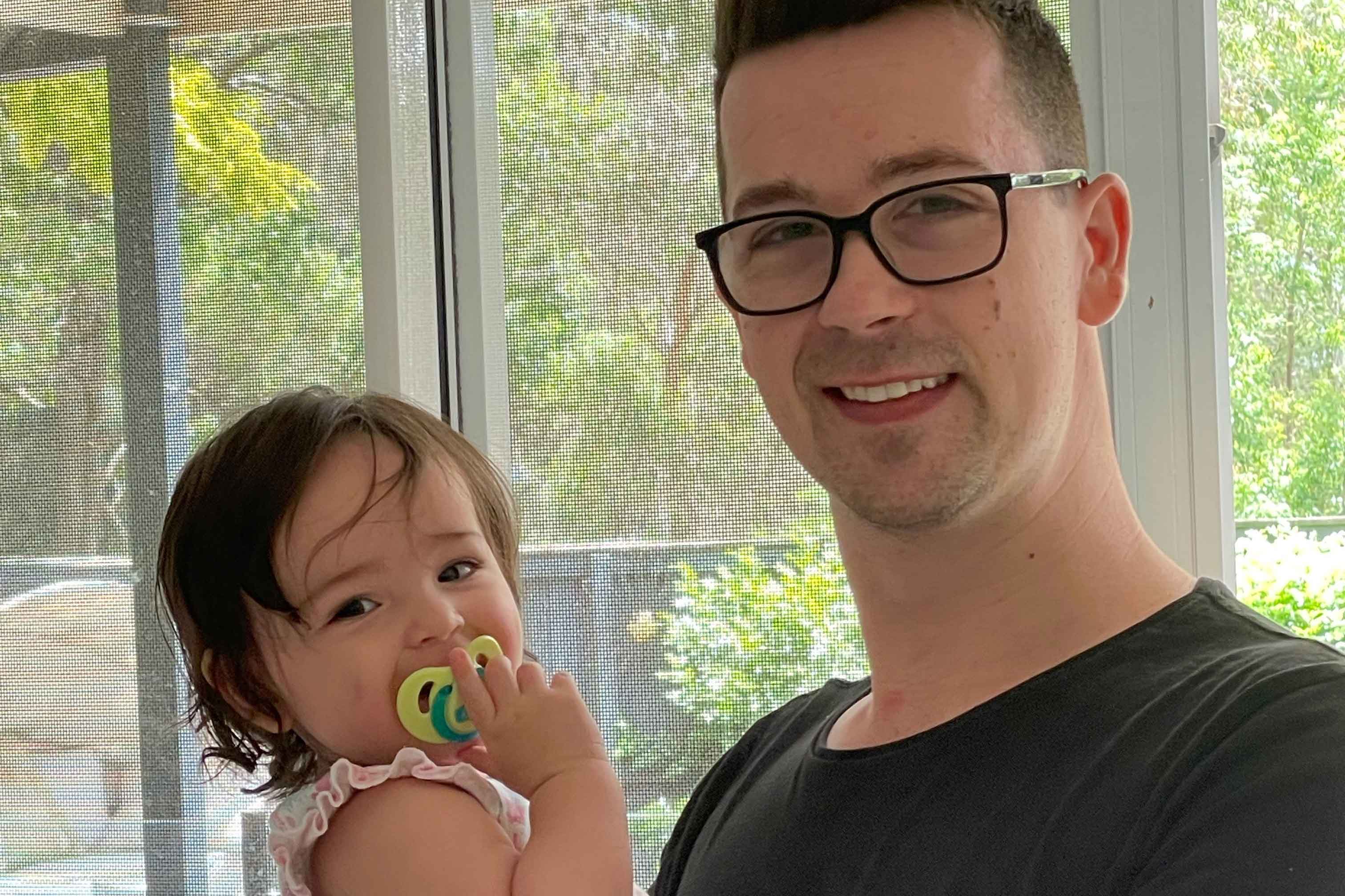 A man holds his young daughter in his arms as they both smile at the camera