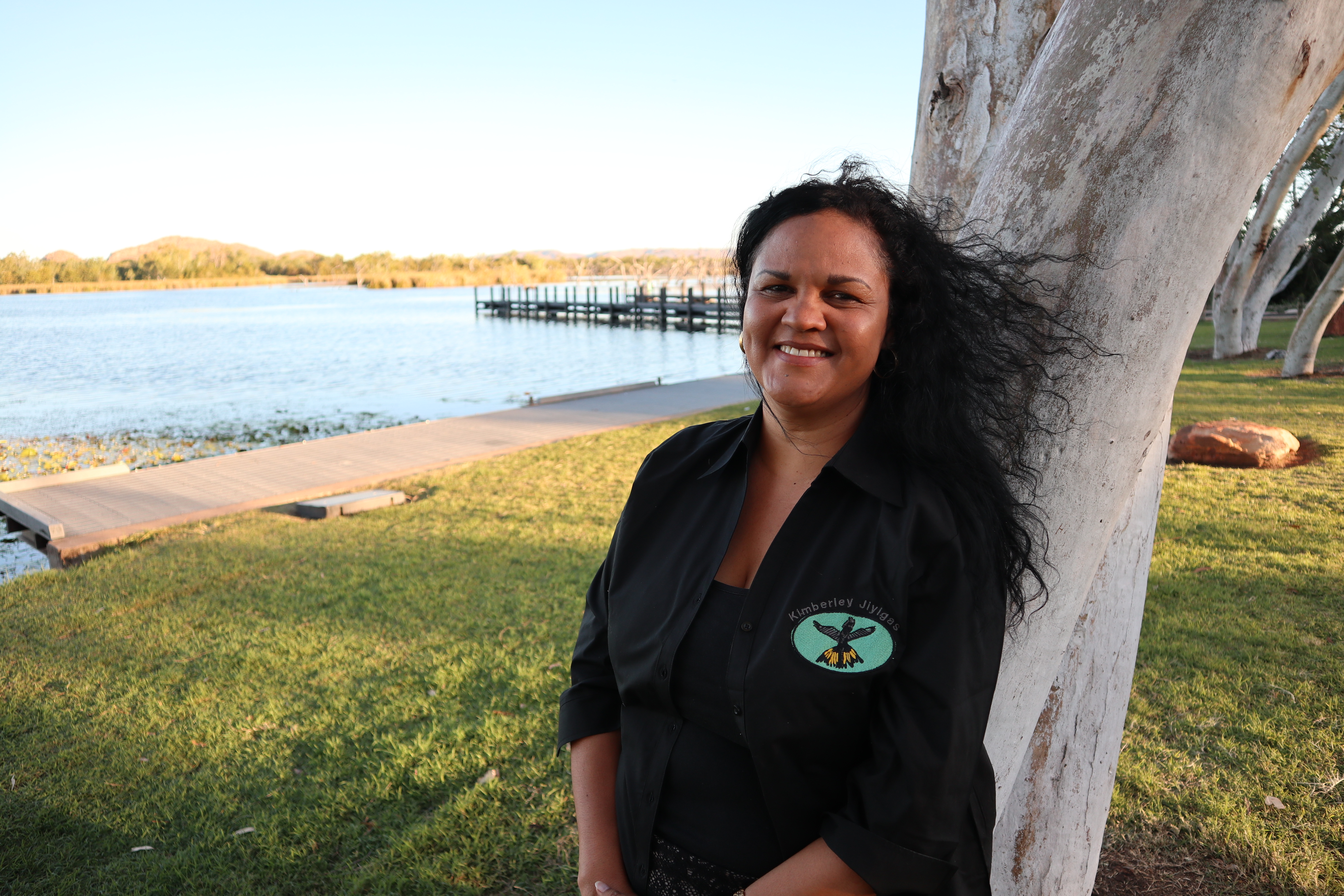 Natasha Short A woman leans against a tree beside a walkway at the water's edge as she smiles at the camera