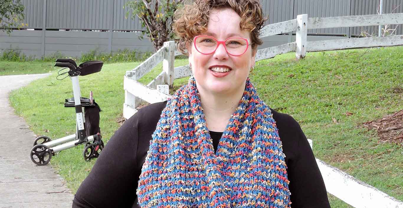 A woman wearing a brightly coloured scarf smiles at the camera as she walks down a concrete footpath using an equally brightly coloured walking stick. Her rollator is on the footpath behind her.