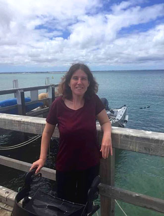 A woman stands beside the water with one arm resting on a railing and the other on her walker