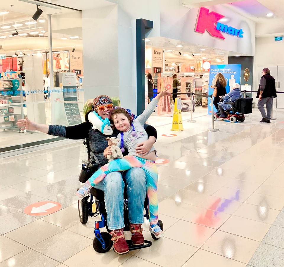 A lady is seated in a wheelchair with a young girl on her knee outside KMart. Both are smiling and the lady has her arms spread out wide. Several people including an older person using a mobility scooter are seen entering the store behind her.