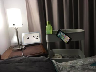 Image show the top of a bedhead beside a bed table with a lamp. Beside this is a tiered trolley with a mobile phone attached to its side on an angle