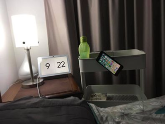 Sarah Wellington Image show the top of a bedhead beside a bed table with a lamp. Beside this is a tiered trolley with a mobile phone attached to its side on an angle
